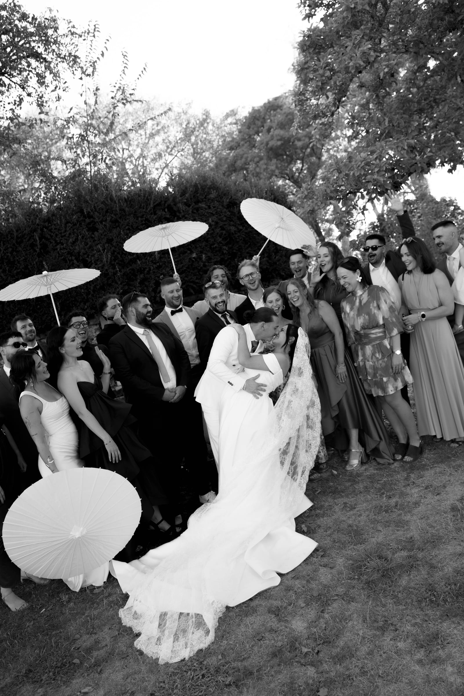 En noir et blanc, le marié renverse la mariée pour un baiser devant leurs invités hilares, ombrelles blanches levées.