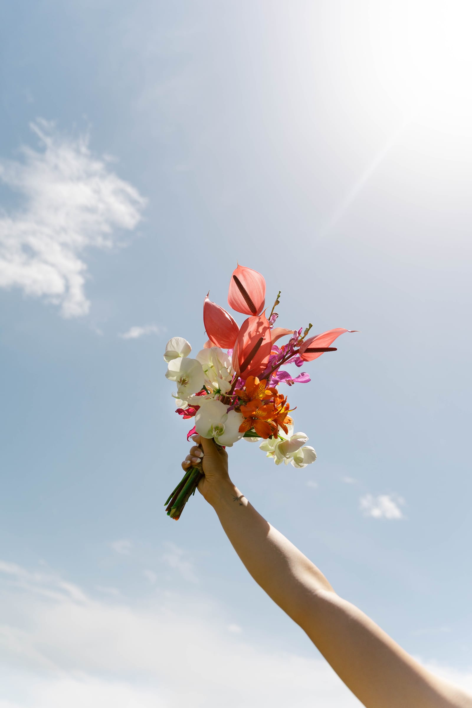 Un bras levé vers le ciel brandit un bouquet d’anthuriums roses, d’orchidées blanches et de fleurs orangées.