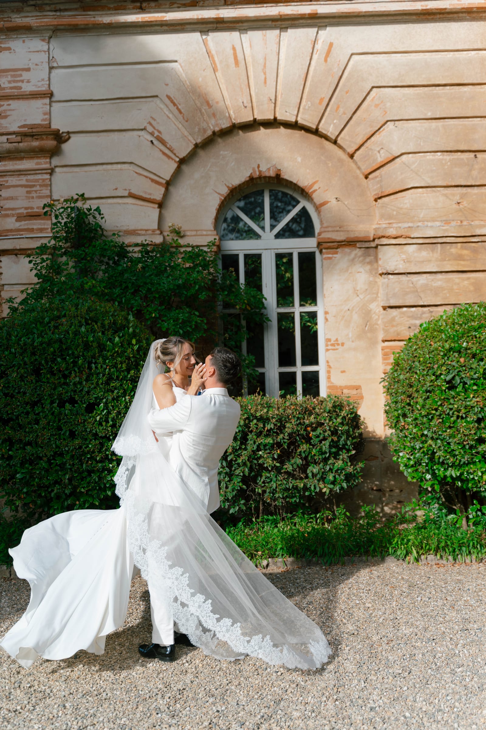 Devant une façade de brique et de pierre, le marié soulève la mariée, son long voile de dentelle déployé.