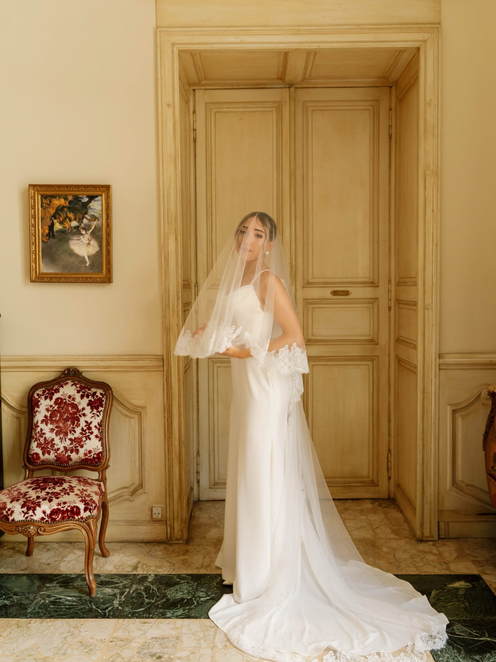 La mariée, voile sur la tête, devant une porte à moulures dans un salon aux boiseries jaunes, près d’un fauteuil ancien à motifs rouges et d’un tableau encadré.