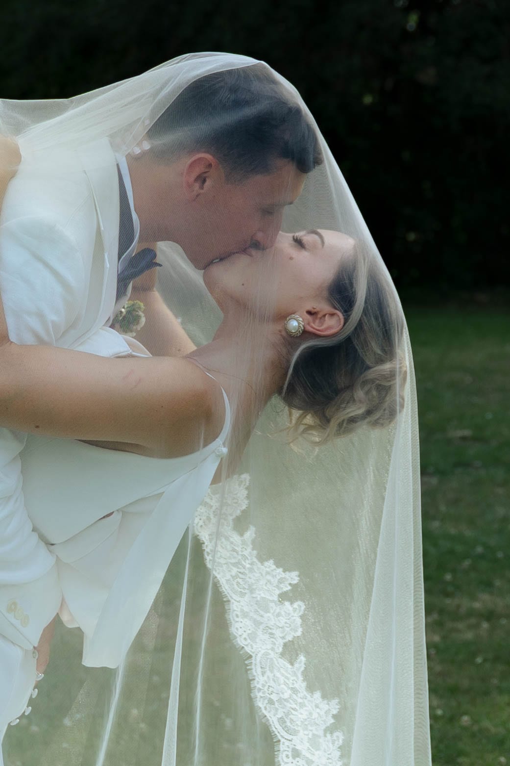 Les mariés, tous deux vêtus de blanc, s’embrassent sous le voile, penchés au-dessus d’une pelouse.