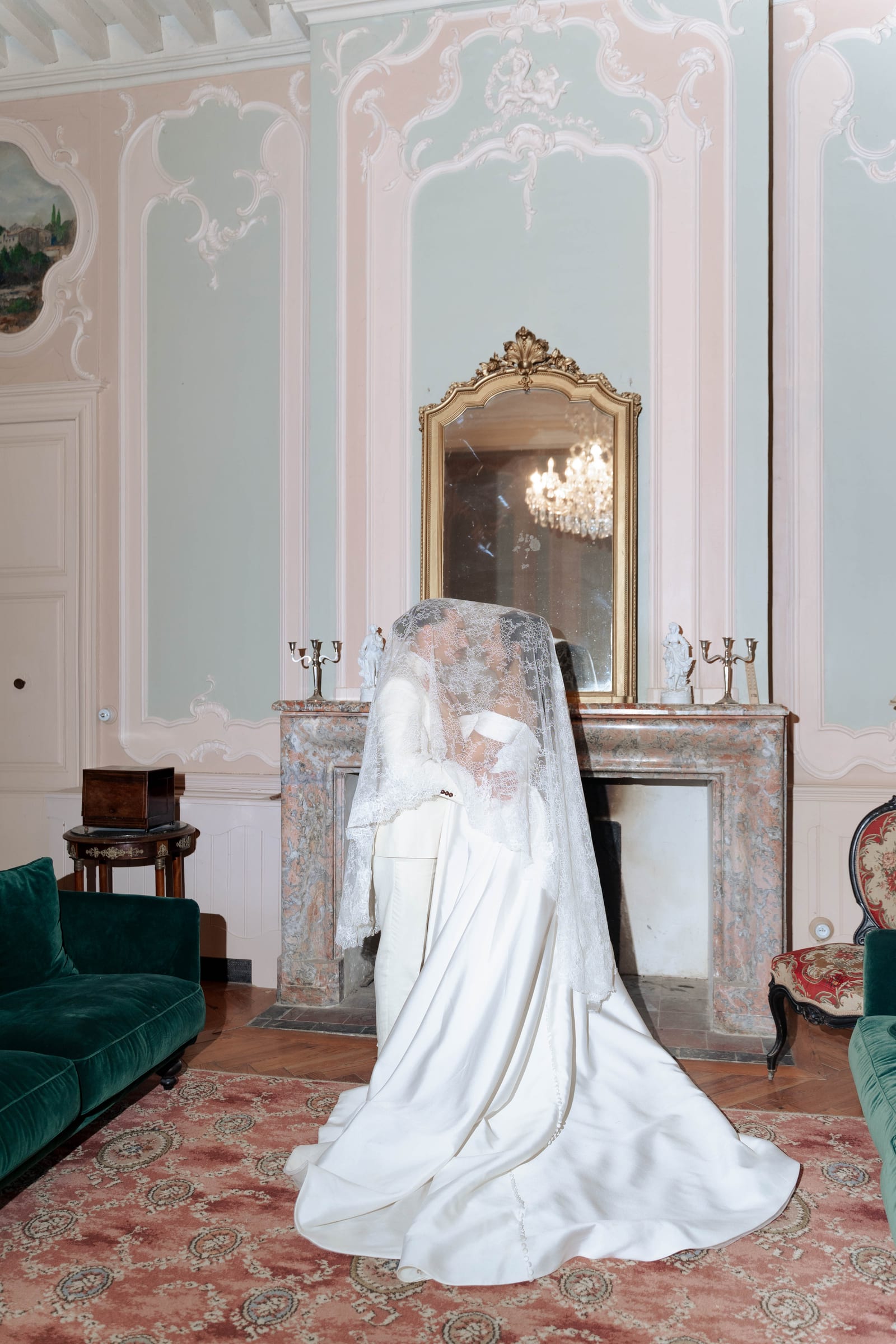 Les mariés enlacés sous un long voile de dentelle devant la cheminée de marbre d’un salon haussmannien, entre canapés vert canard et miroir doré.